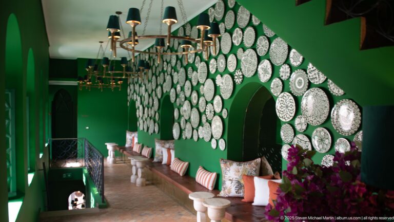 Long green interior with brass chandeliers, built-in bench seating, cushions, white tables, flowers, arches, and a wall of patterned plates.