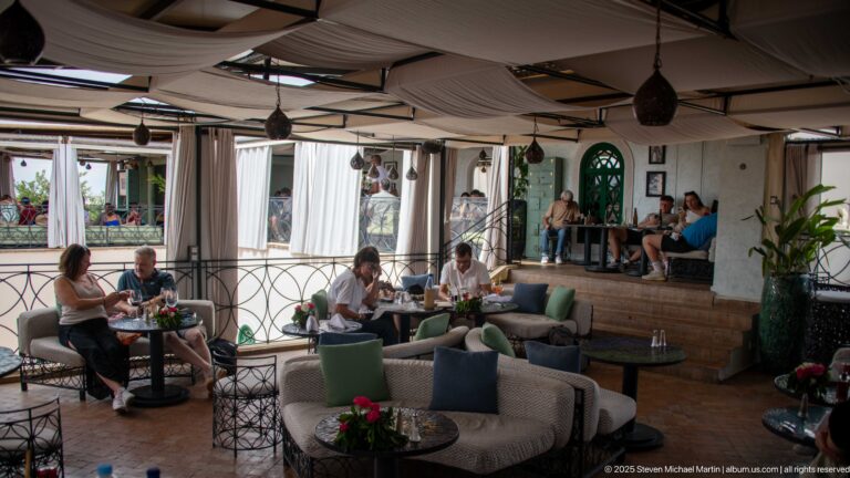 Covered rooftop café lounge with sofas, small tables, white curtains, hanging lamps, diners, and potted plants.