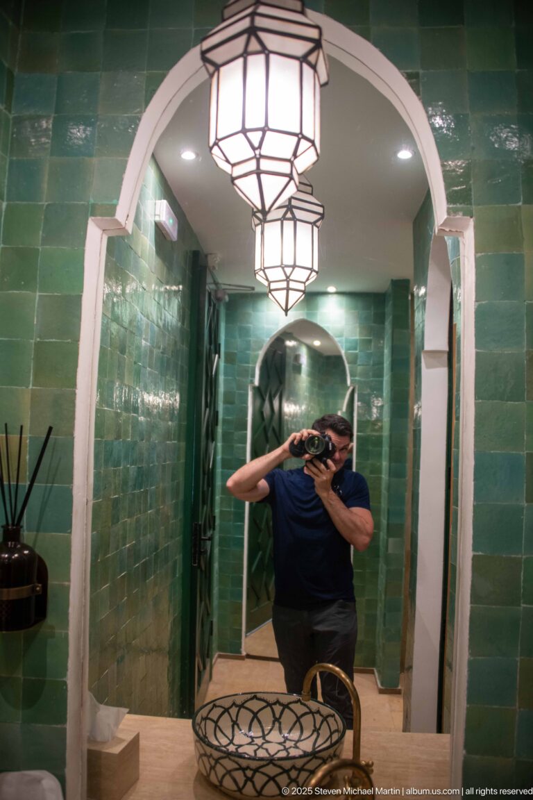Photographer reflected in a mirror inside a narrow green-tiled washroom with white arches and geometric hanging lights.
