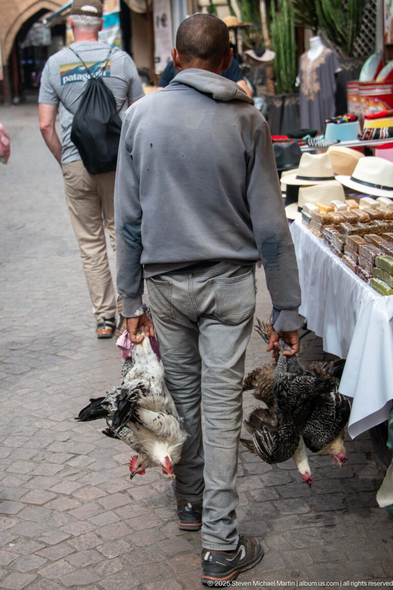 Man carrying several slaughtered chickens by their legs through a busy Marrakech market lane beside food stalls.
