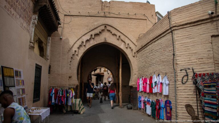 Large pointed arch through earthen-colored walls with clothing and textile stalls flanking a busy market passage.