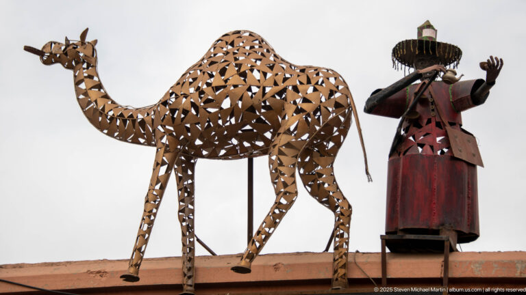 Openwork metal camel beside a stylized musician sculpture mounted above a Marrakech roofline.