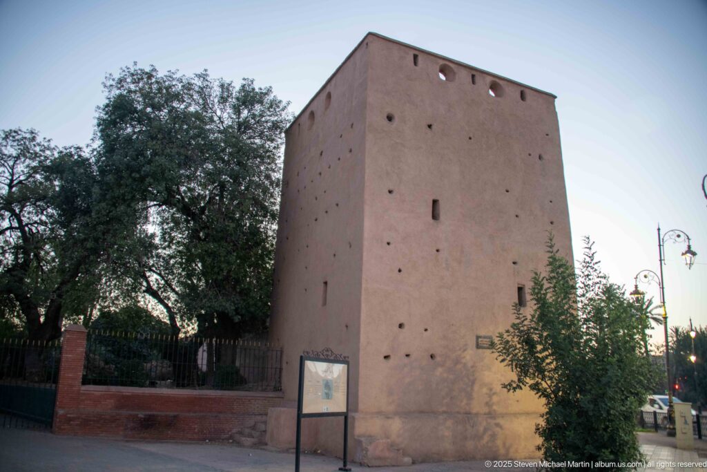 Tall square pink-ochre defensive tower with small openings beside trees and a landscaped roadside area in Marrakech.