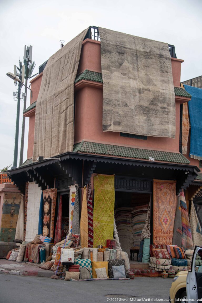 Roadside Marrakech carpet shop with large rugs hanging from the façade and rolled textiles and cushions stacked below.