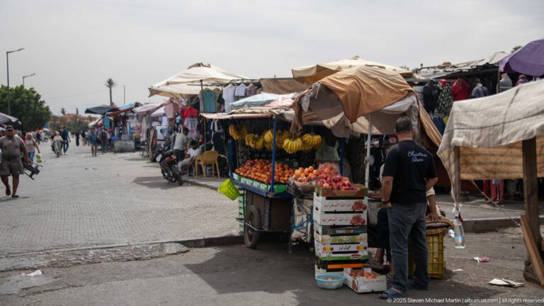 Open-air Marrakech produce stall with bananas, tomatoes, crates, boxes, canvas awnings, shoppers, and a broad market lane.
