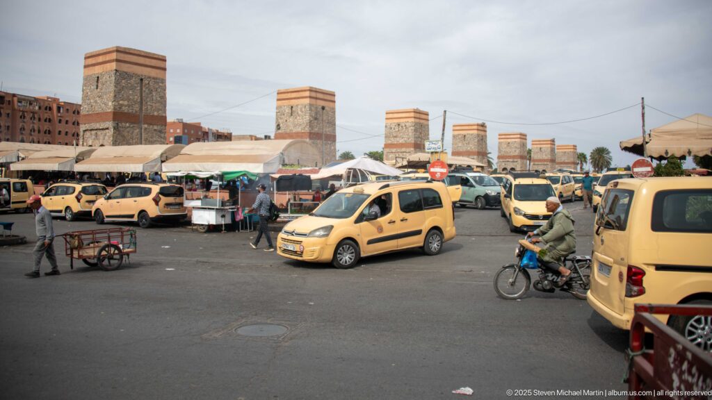 Busy Marrakech intersection with beige petit taxis, a motorbike, handcart, market awnings, and square towers in the distance.