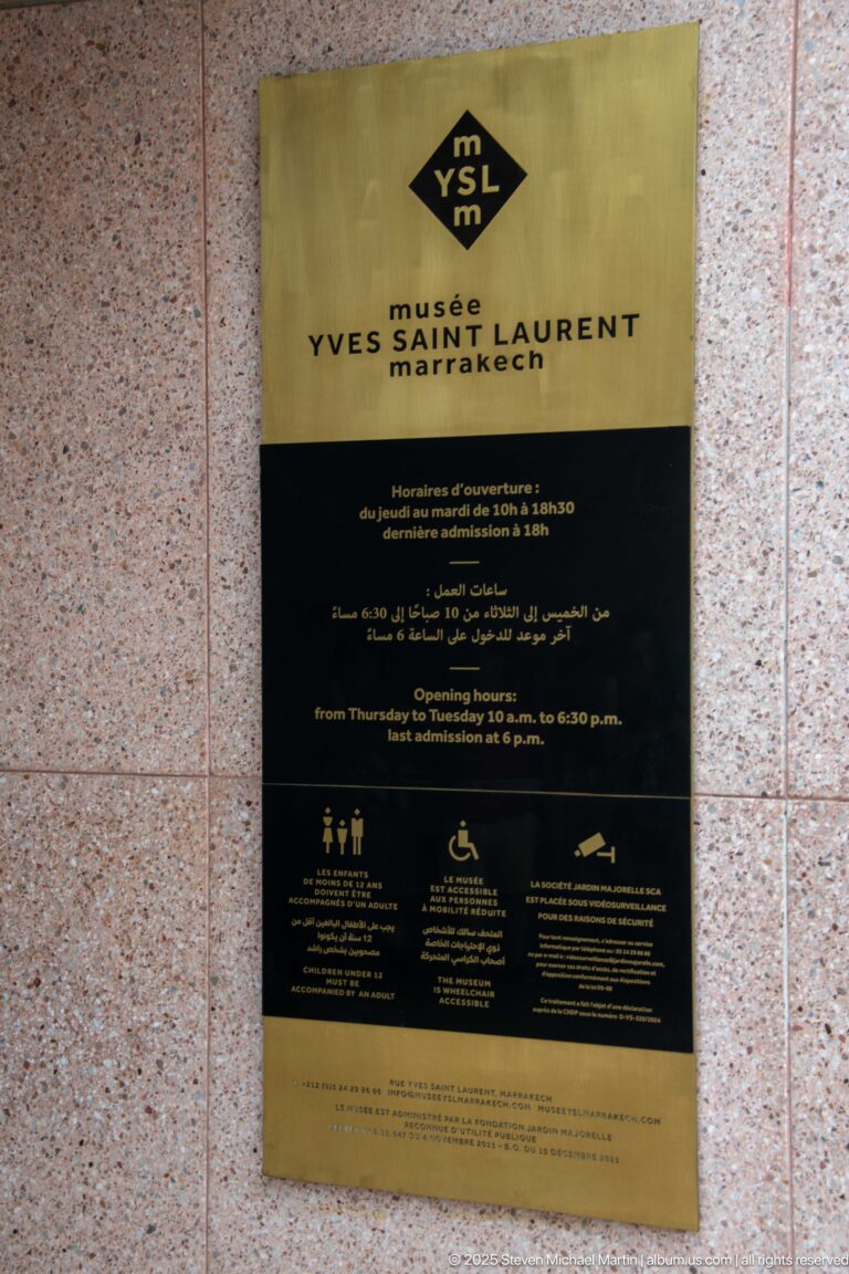 Brass and black Musée Yves Saint Laurent Marrakech information plaque with YSL logo, opening hours, multilingual text, and visitor icons.