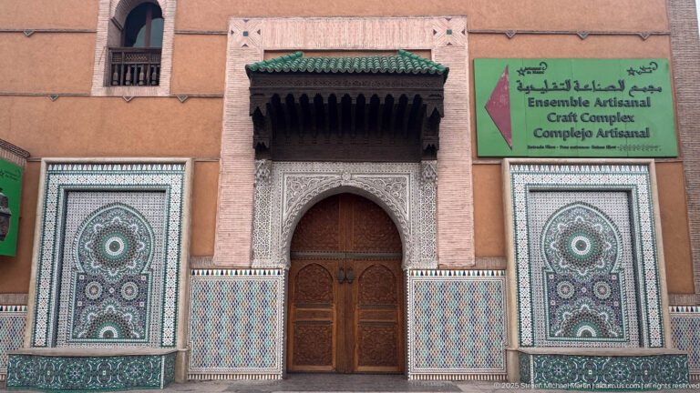 Ensemble Artisanal entrance with carved plaster, zellij panels, green-tiled canopy, wooden door, and multilingual craft-complex sign.