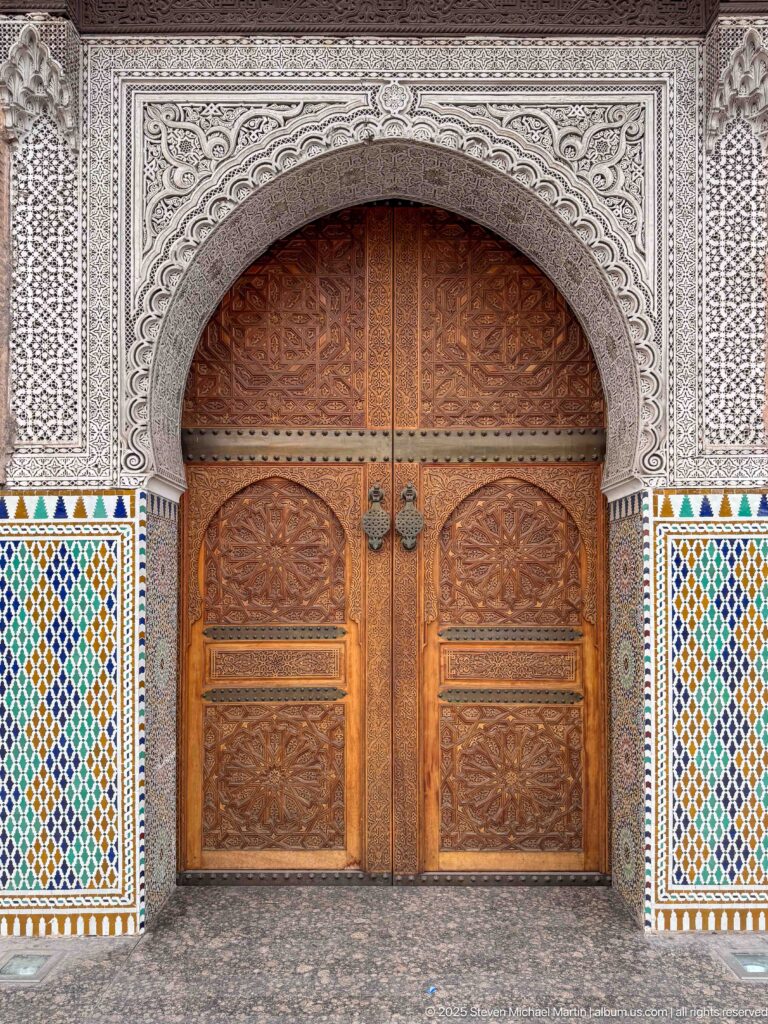 Carved wooden double doors beneath an ornate horseshoe arch with white plaster carving and multicolored zellij panels.