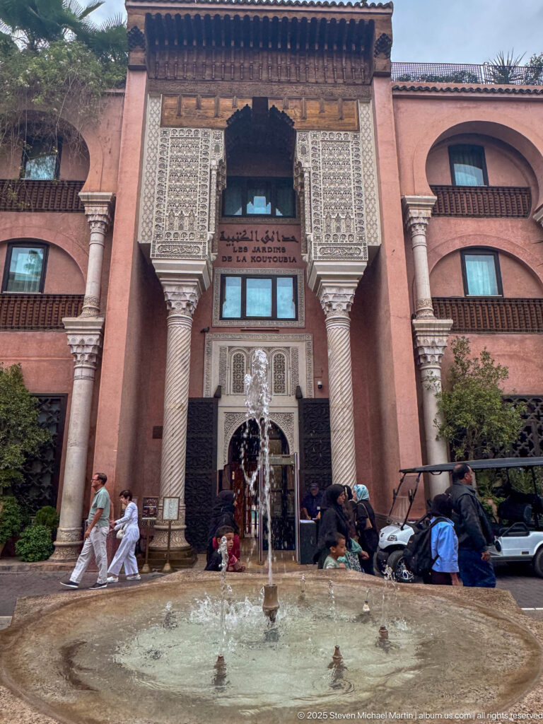 Ornate pink entrance with fountain, tall columns, carved plaster panels, dark timber eaves, and Arabic signage above the doorway.