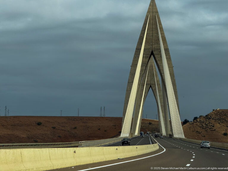Modern cable-stayed highway bridge with tall pointed concrete A-frame pylons beneath a dark cloudy sky.