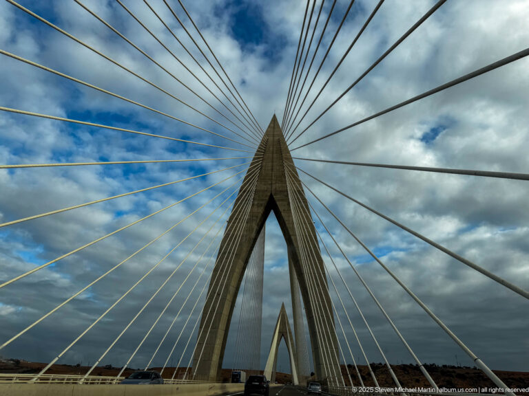 Upward view of a pointed concrete bridge pylon with radiating stay cables and a second pylon visible through the opening.