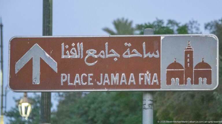 Brown Arabic and Latin-script sign pointing straight ahead to Place Jamaa Fna, with a mosque pictogram.