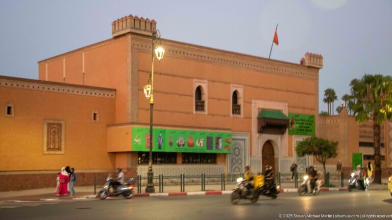 Large pink public building with arched windows, banners, Moroccan flag, palms, scooters, and pedestrians at dusk in Marrakech.