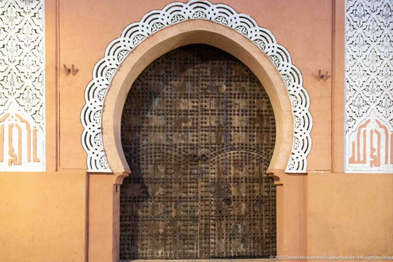 Tall dark wooden door inside a horseshoe arch framed by white geometric decoration on a pink Marrakech façade.