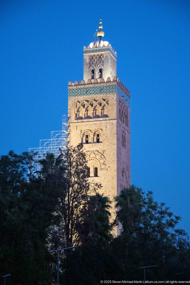 Illuminated Koutoubia Mosque minaret above dark trees at blue hour, with scaffolding visible along one side.