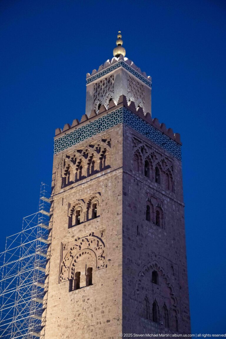 Close view of the illuminated upper Koutoubia minaret with arched decoration, green-tiled band, finial, and side scaffolding.