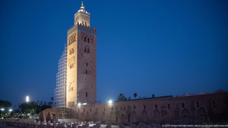 Illuminated Koutoubia minaret with scaffolding rising above the long mosque exterior wall at night in Marrakech.