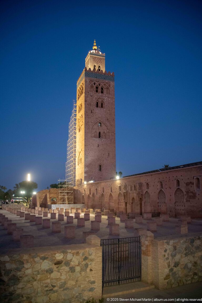 Koutoubia minaret at night beyond low masonry, paved grounds, and the arched exterior wall of the mosque precinct.