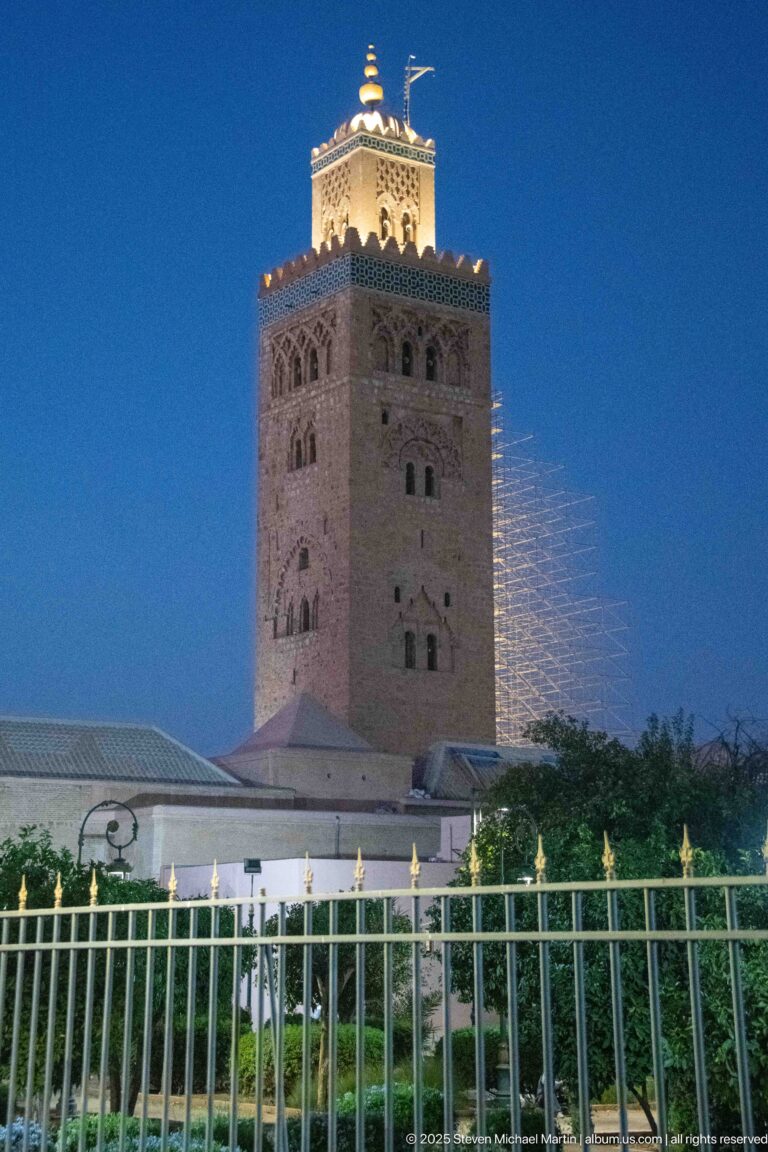 Illuminated Koutoubia minaret with side scaffolding seen beyond trees, low walls, and a metal fence at night.