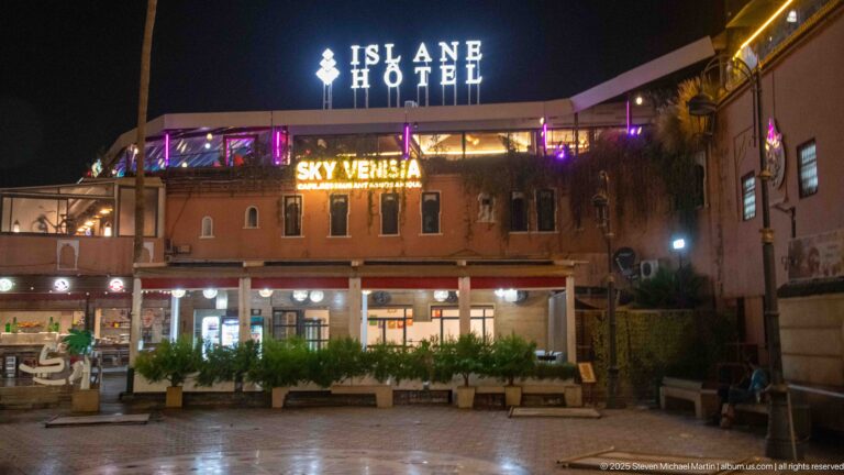 Islane Hotel at night with bright ISLANE HOTEL and SKY VENISIA signs above terraces and street-level businesses.
