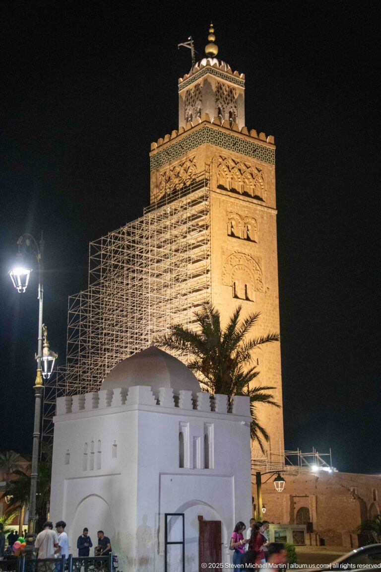 Illuminated Koutoubia minaret wrapped partly in scaffolding behind a low white pavilion at night in Marrakech.