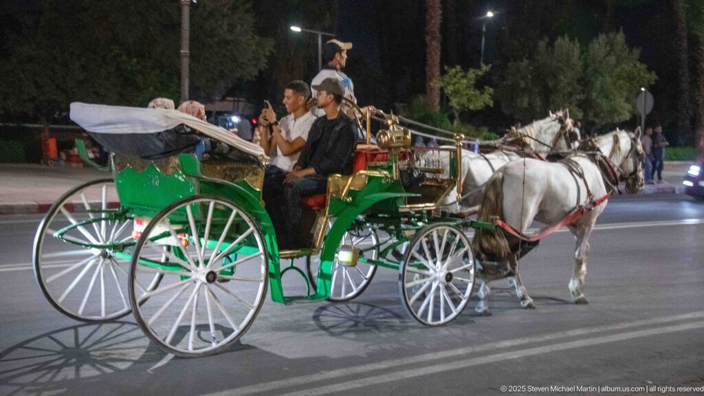 Green horse-drawn calèche carrying passengers through a lit Marrakech street at night, pulled by two pale horses.