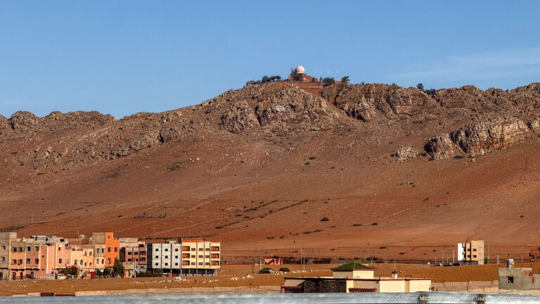 Low apartment blocks below a barren reddish-brown rocky ridge under a clear blue sky on the outskirts of Marrakech.