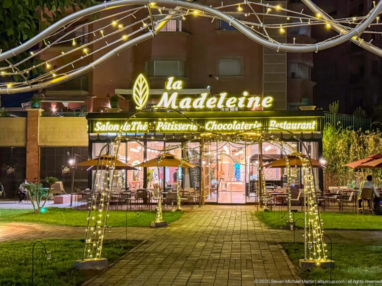 Illuminated La Madeleine restaurant entrance at night with decorative light arches, glass storefront, outdoor seating, and landscaped paving.