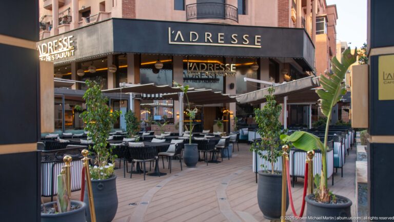 L’Adresse restaurant terrace with shaded tables, black façade, potted trees, and decorative planters along a Marrakech street.