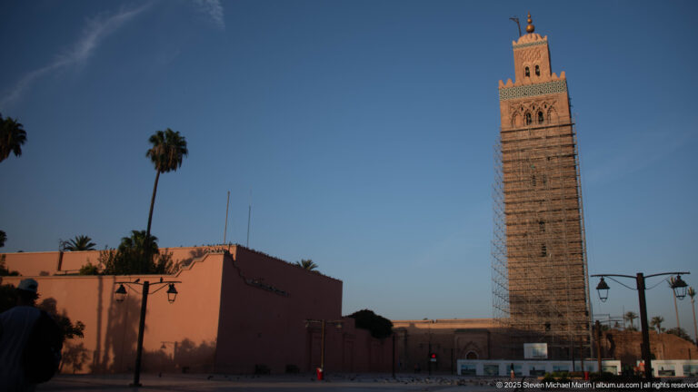 Koutoubia Mosque minaret above low pink buildings and palms, with partial scaffolding visible against a blue sky.