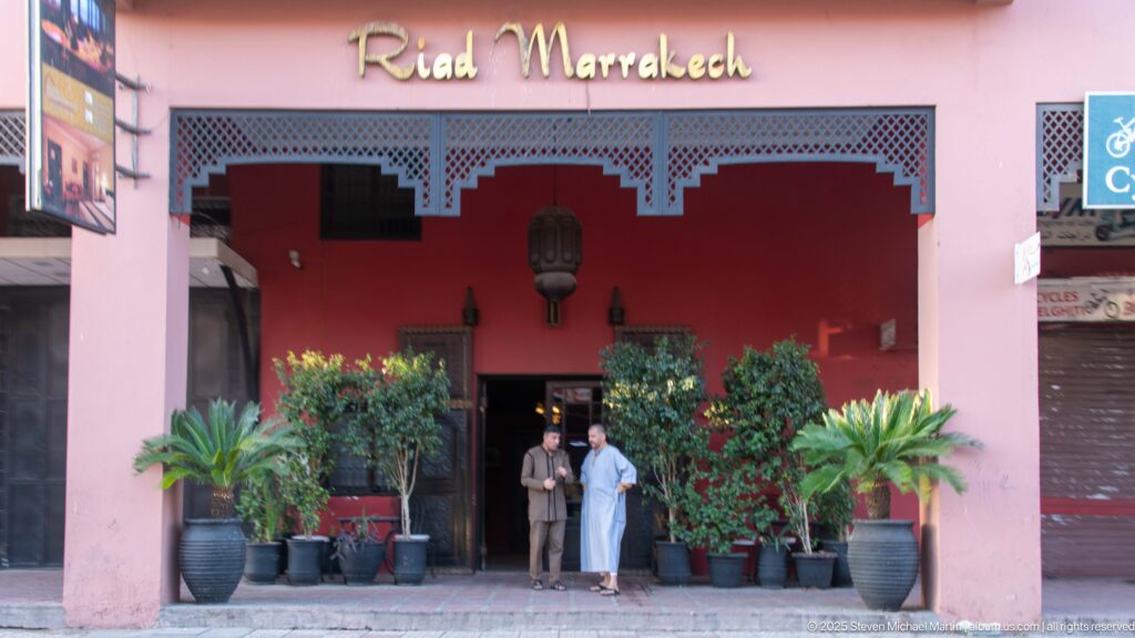 Pink Riad Marrakech entrance with red recessed doorway, decorative edging, potted plants, and two people standing outside.