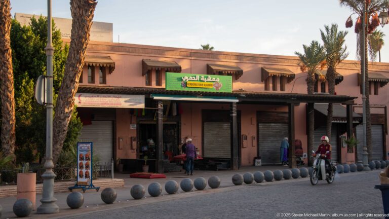 Low pink commercial buildings with awnings, small shops, palm trees, pedestrians, and spherical street bollards in Marrakech.