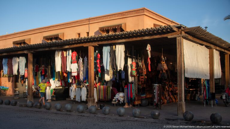 Open-fronted Marrakech market stall with hanging clothing, textiles, bags, and leather goods beneath a timber pergola.