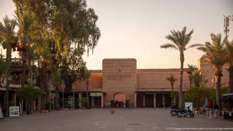 Arched gateway in massive pink walls across an open plaza with palm trees and a few parked motorcycles.