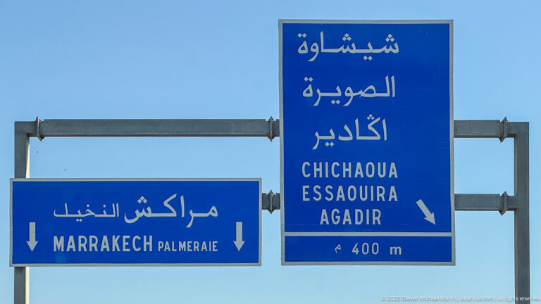 Blue Arabic and Latin-script road signs point toward Marrakech Palmeraie, Chichaoua, Essaouira, and Agadir at an upcoming junction.