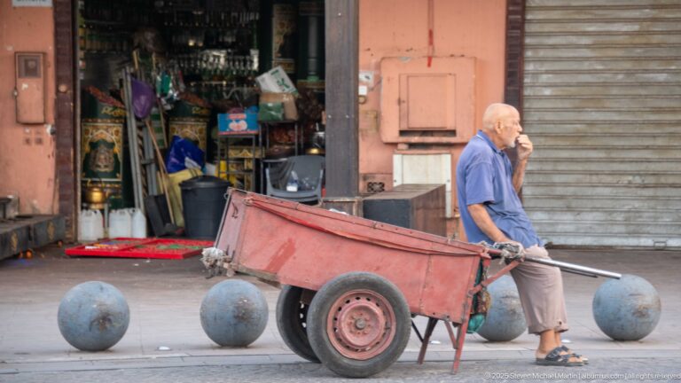 Older man pulling a red two-wheeled handcart past a small shop and spherical bollards in Marrakech.