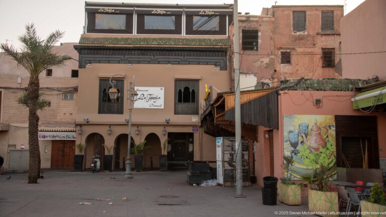 Pink multi-story commercial building with arched ground-floor openings, shuttered windows, a palm tree, and neighboring weathered walls.