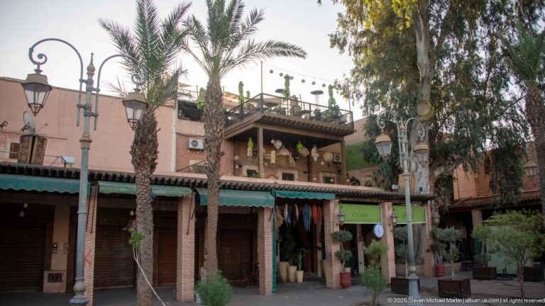 Palm-lined Marrakech shopfronts below a timber-shaded upper terrace, with potted plants and traditional-style street lamps.