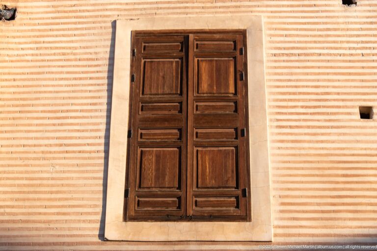 Closed dark wooden shutters set in a rectangular opening within a horizontally striped pale and warm-toned masonry wall.