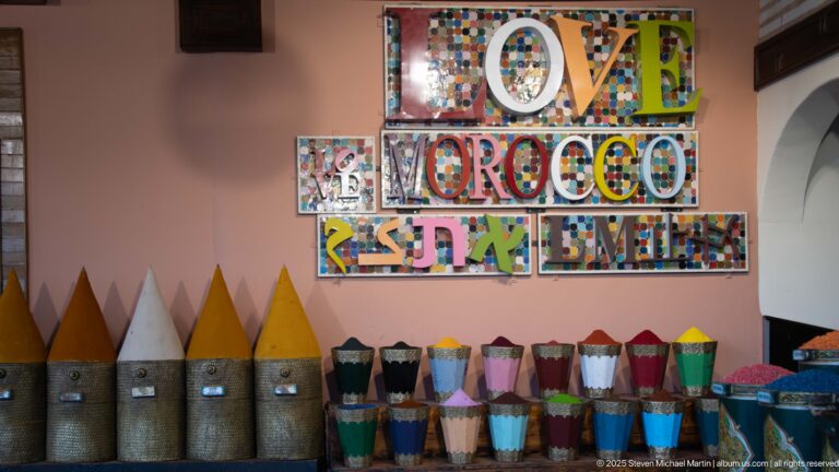 Colorful mosaic-letter “LOVE MOROCCO” sign above rows of open containers filled with brightly colored powders.