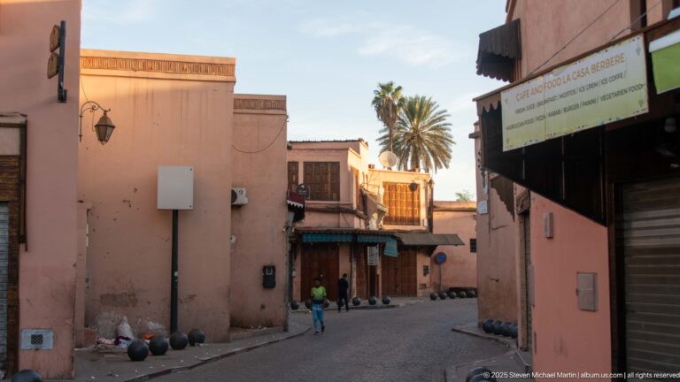 Narrow Marrakech street between pink walls, with palms, distant pedestrians, shopfronts, and spherical bollards.