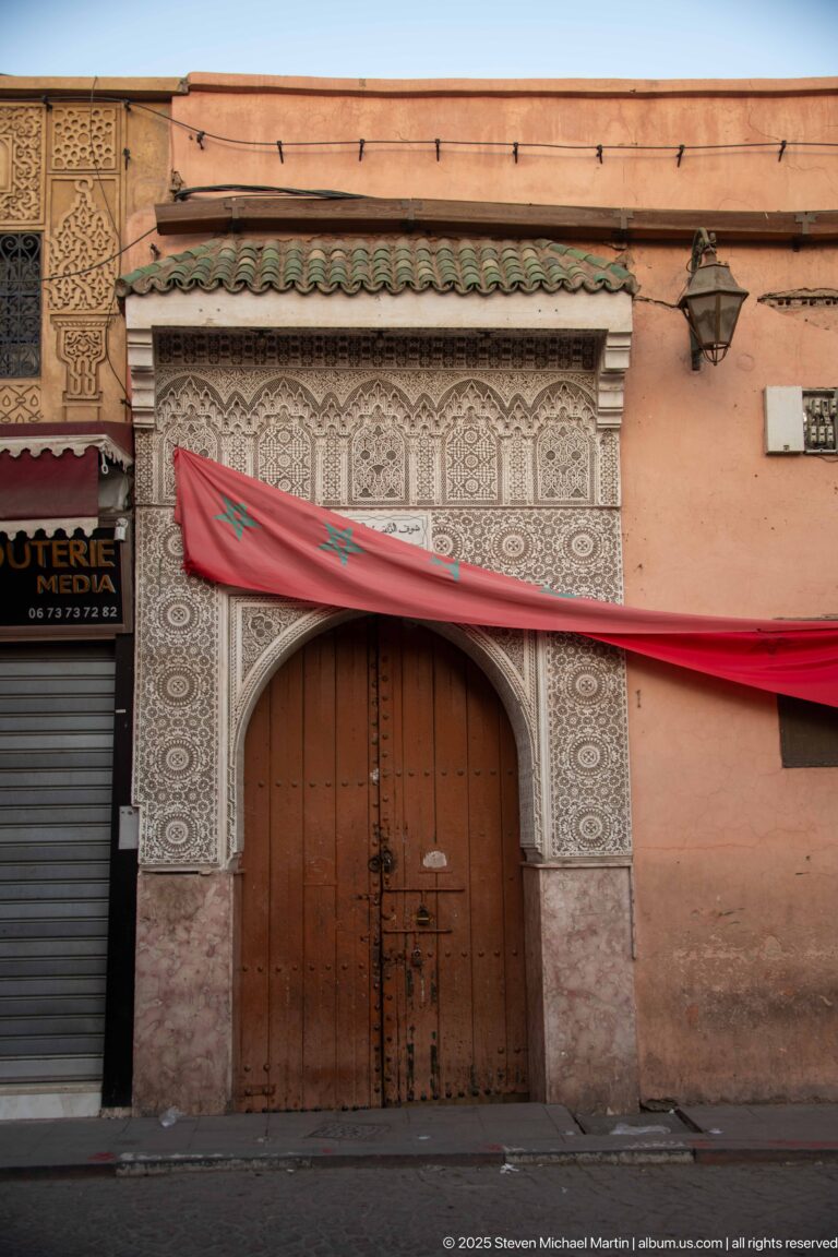 Large wooden double door in an ornate carved surround with green tile cap and Moroccan flag above.
