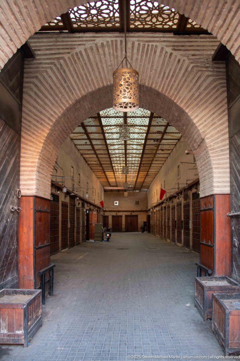 Wide brick arch opening into a roofed Marrakech market passage with closed stalls, overhead latticework, and hanging lanterns.