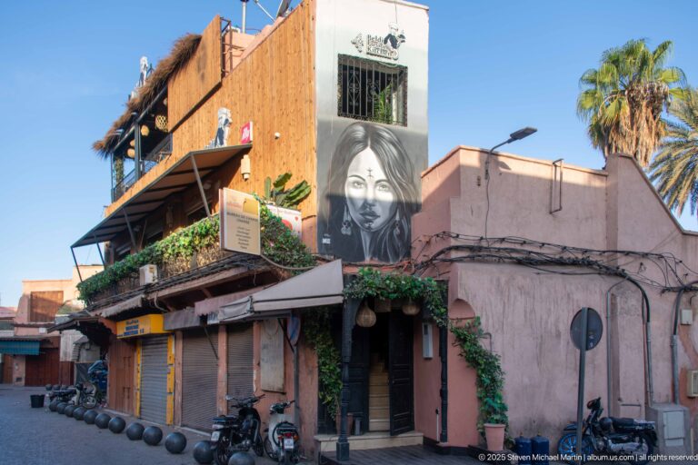 Large black-and-white portrait mural on a pink corner building above closed shops and parked motorbikes in Marrakech.