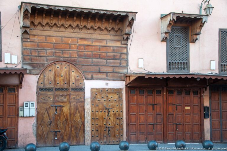 Three varied wooden doorways in a pink wall, including arched studded doors, paneled doors, timber eaves, and an upper screened window.