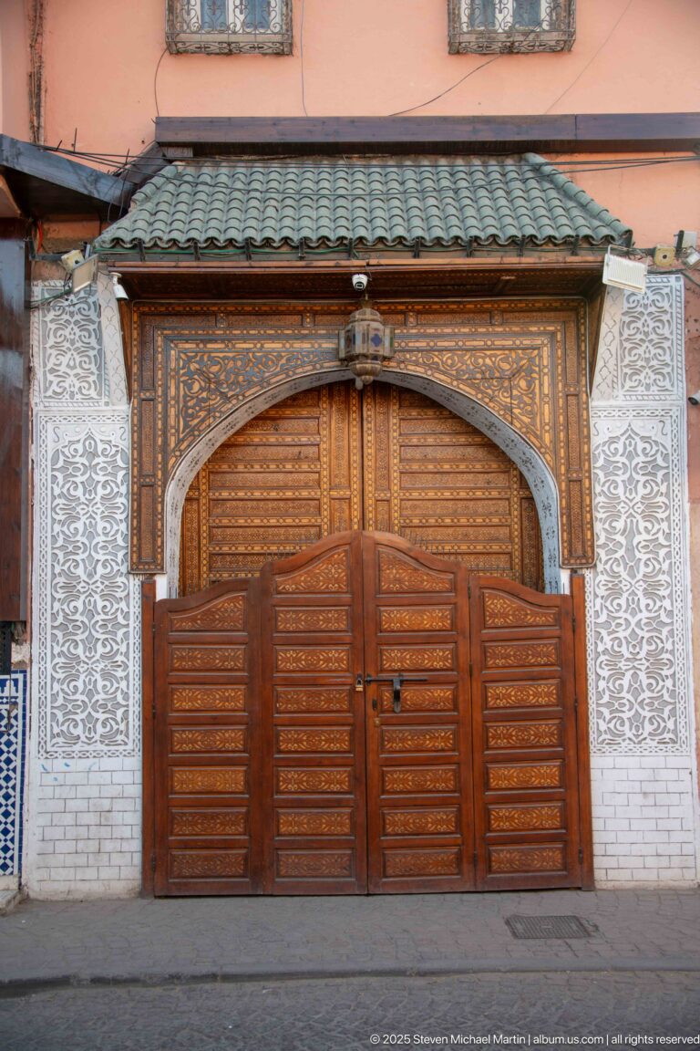 Large carved wooden portal with ornate upper arch, white patterned wall relief, and a green-tiled canopy.