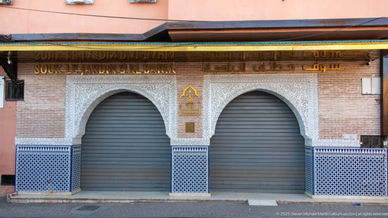 Two closed horseshoe-arched shop openings with carved white ornament, blue geometric tilework, brickwork, and a wooden canopy.