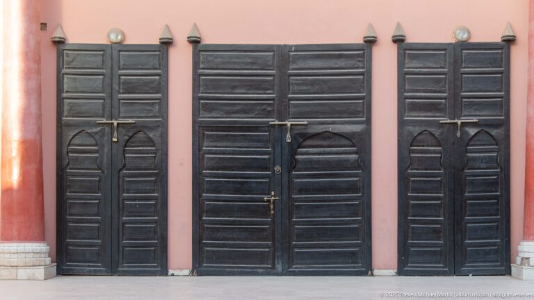 Triple black wooden doorway with paneled leaves, metal latches, pointed lower motifs, and a surrounding pink wall.