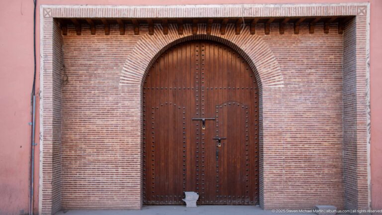 Large rounded wooden gate with metal studs and central latch set within a broad brick arch in Marrakech.